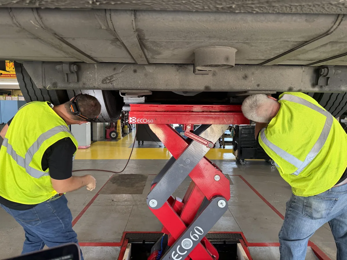 Two Traveling Wrenches technicians performing an underbody inspection on a bus raised on an in-ground lift